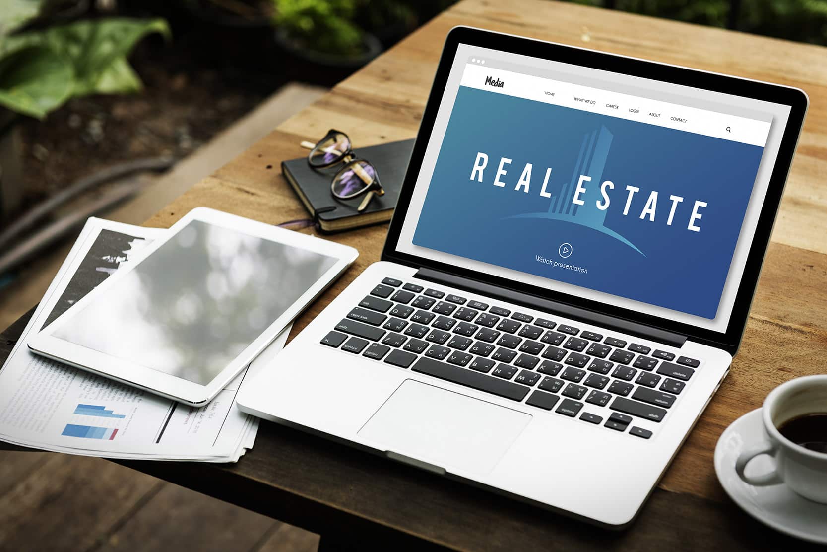 A computer on a desk with "Real Estate" in the screen. Tablet, glasses and a cup of coffee also on table